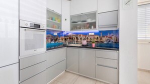 A modern kitchen with white cabinets, a built-in oven, and a backsplash depicting a coastal scene. - Apartment Krempy - Two Bedroom Apartment with Terrace (Trogir)