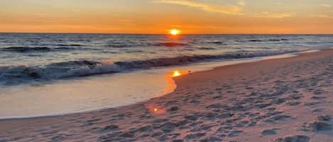 Una playa con huellas que conducen al agua, un atardecer al fondo y olas tranquilas.
