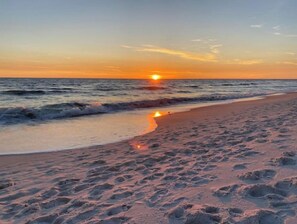 En strand med fodaftryk, der fører ned mod vandet, og en solnedgang i baggrunden samt rolige bølger.