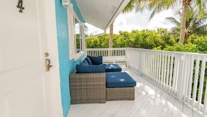 A covered outdoor seating area with wicker furniture and cushions, a white railing, and a view of lush greenery. - Surf Views 2BR - Sept Discounts Now Live! (Fort Pierce)