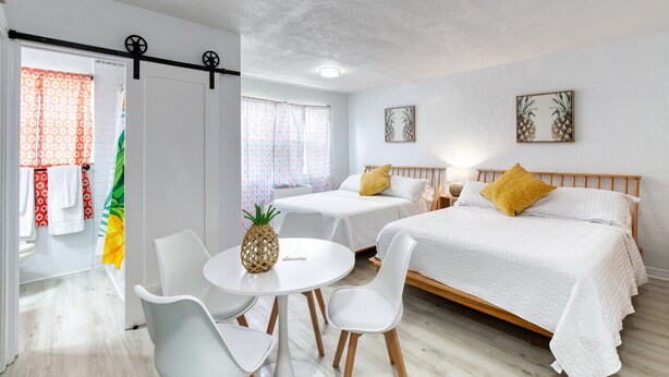 A hotel room with two beds, a small table with chairs, a pineapple on the table, and a view of the beach through the window. - Pineapple Shores Inn Unit 1 (Daytona Beach)