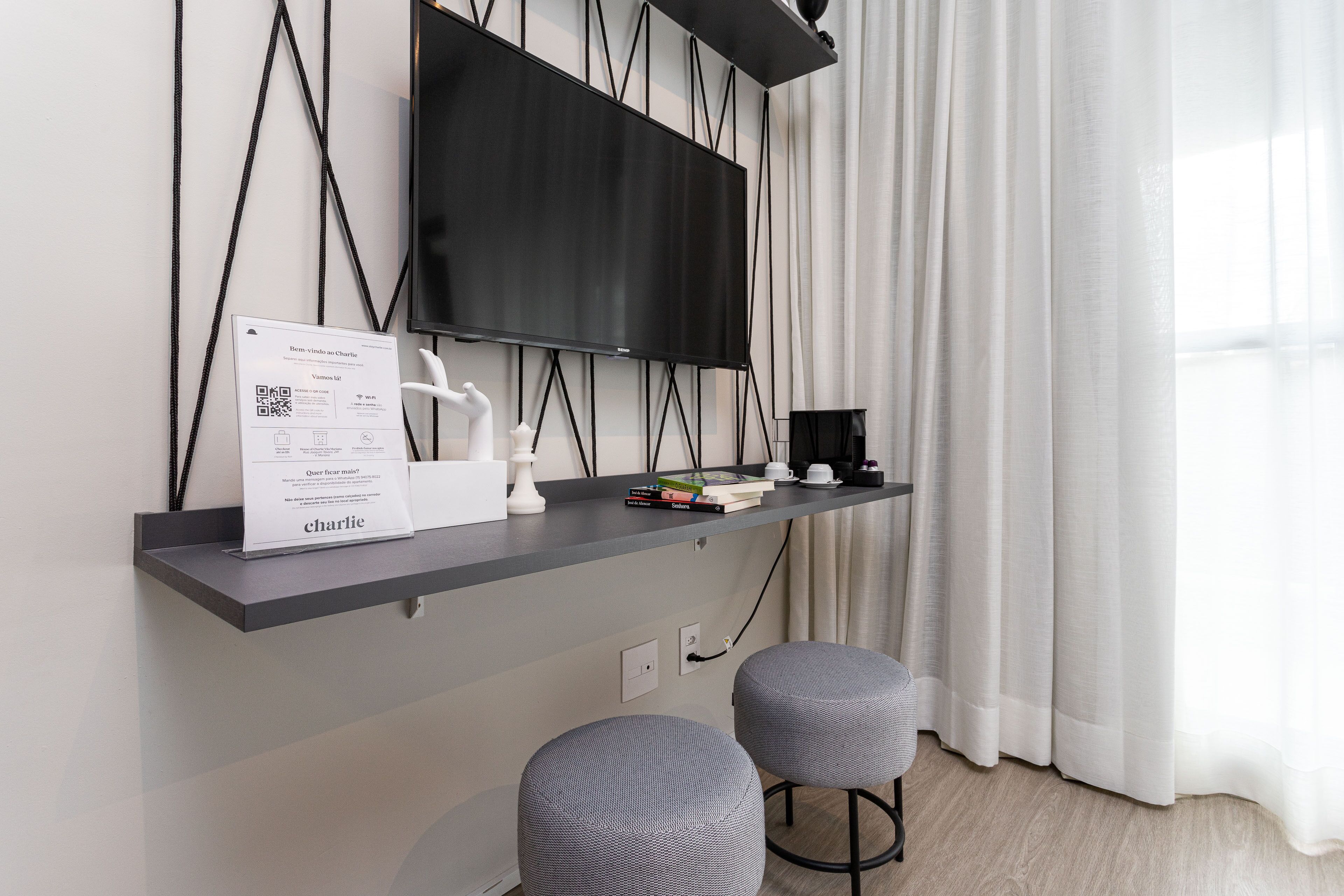 Une chambre moderne avec un téléviseur à écran plat fixé au mur, une petite étagère avec des livres et une tasse, et deux tabourets gris.