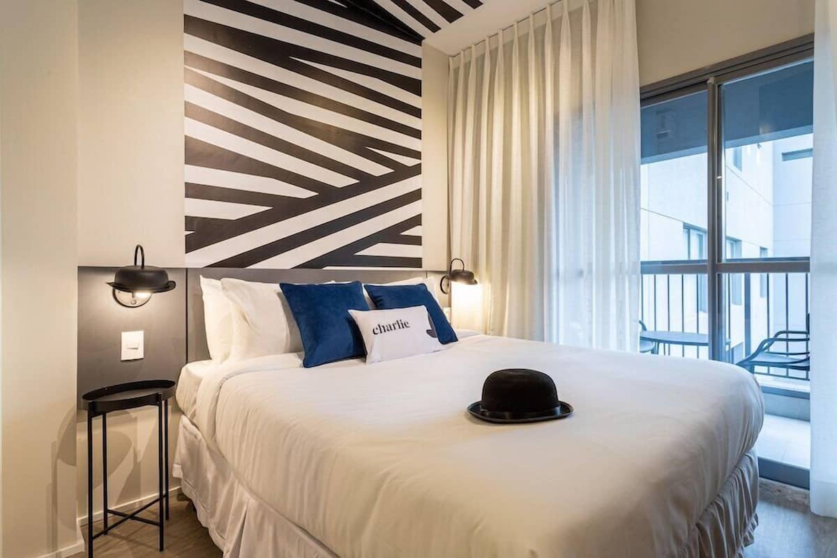 Een moderne hotelkamer met een groot bed, een zwarte hoed op het bed, een muur met een geometrisch patroon en een balkon met een hekwerk.