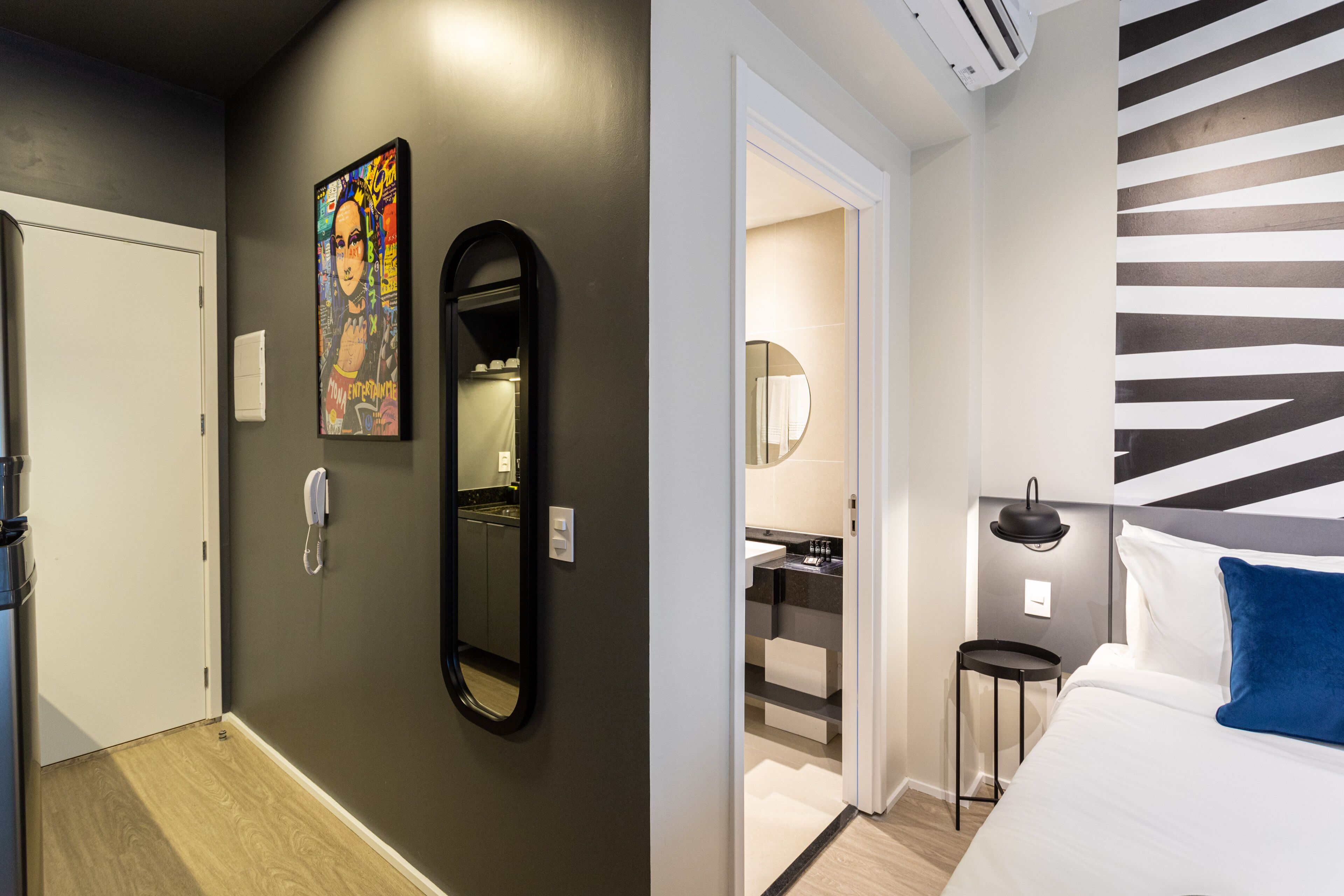 A hotel room with a door, a mirror, a bathroom with a sink, and a bed with a blue pillow.