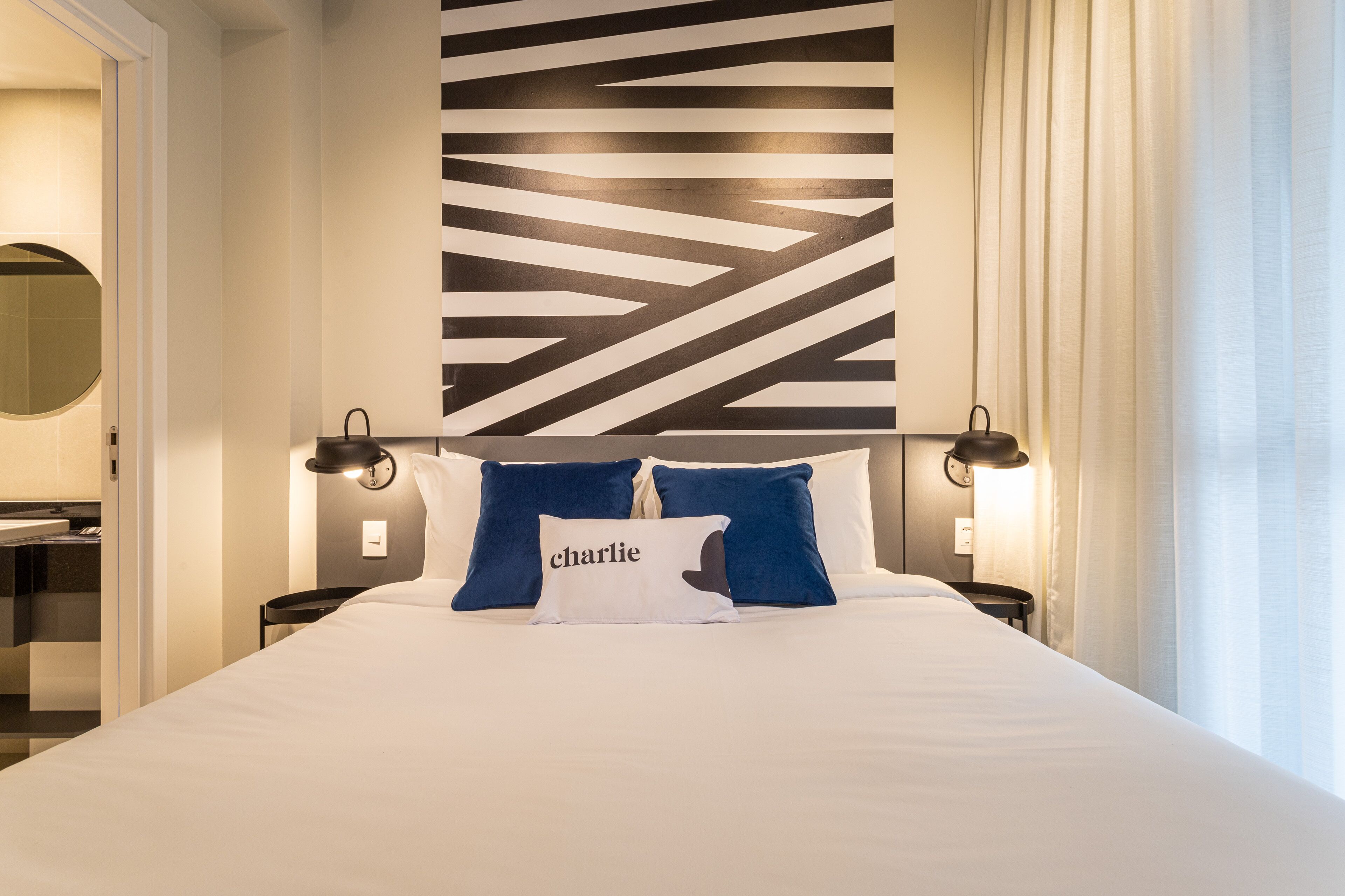Habitación de hotel con una cama grande, dos almohadas, una pared con diseño geométrico, una mesita de noche con lámpara y un espejo.