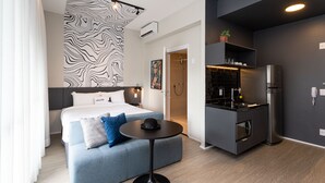 A modern hotel room with a bed, a small table, a kitchenette, and a bathroom.