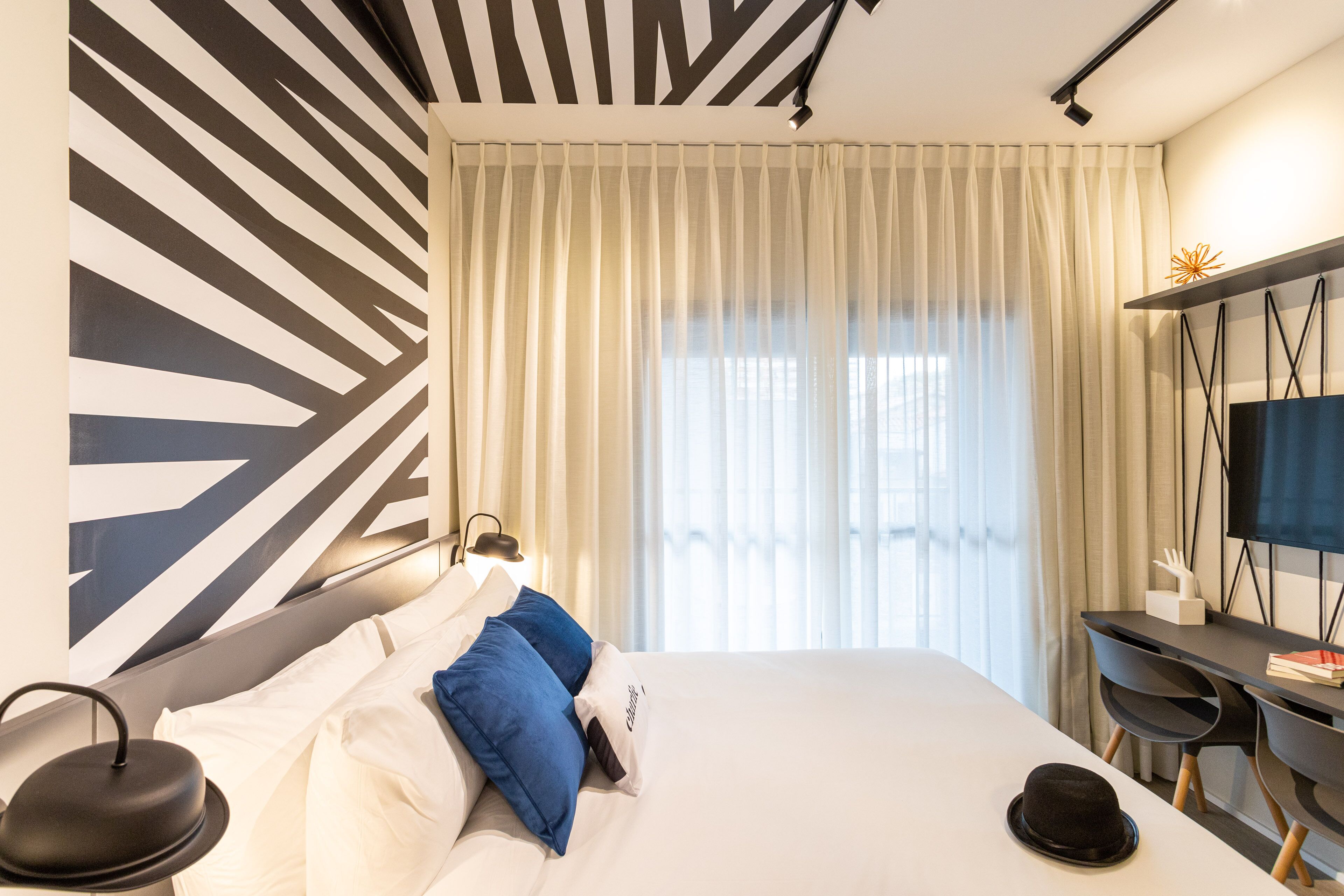 A modern hotel room with a large bed, a desk with a chair, a television, and a decorative wall piece.