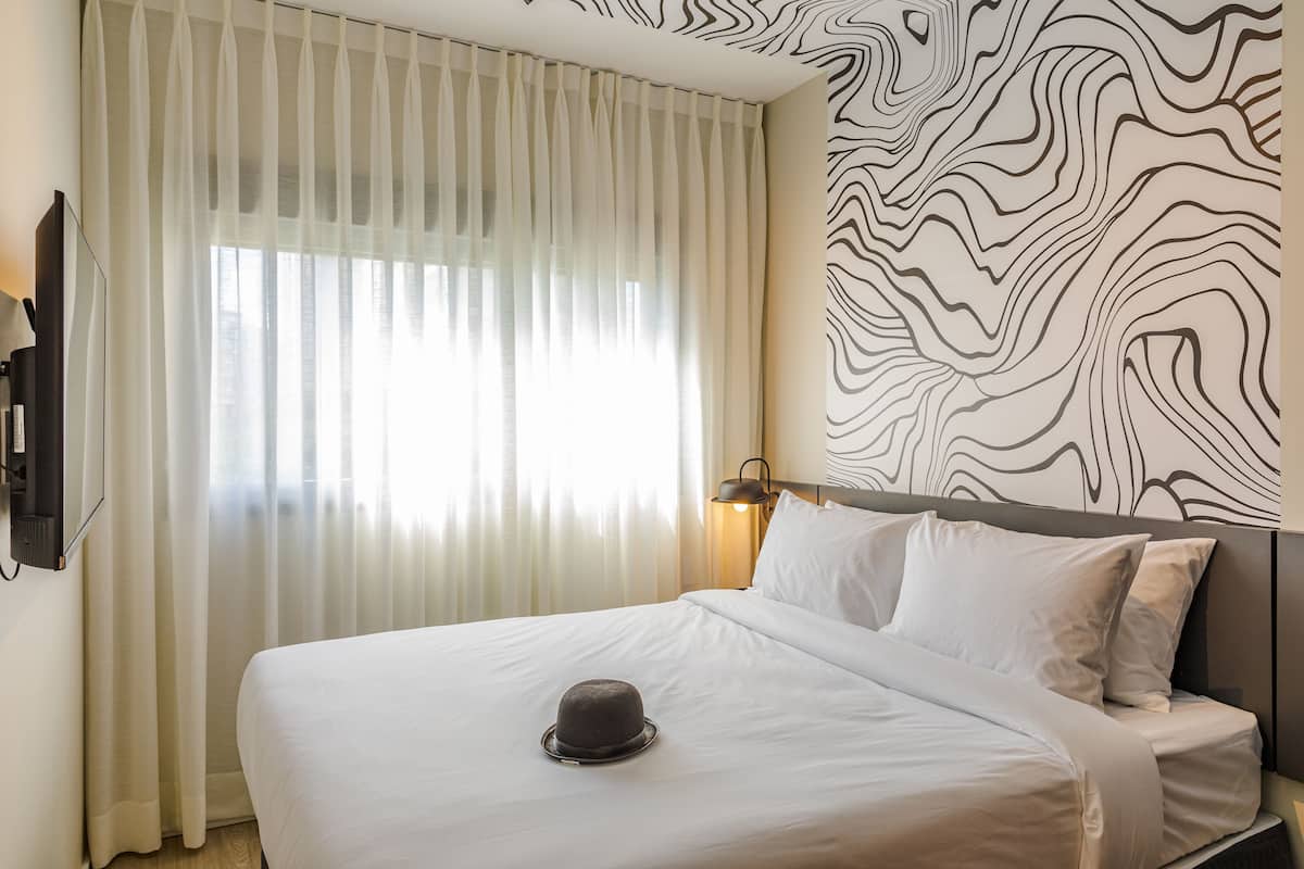 Een hotelkamer met een bed, witte gordijnen, een muur met een zwart-wit abstract ontwerp en een lamp.