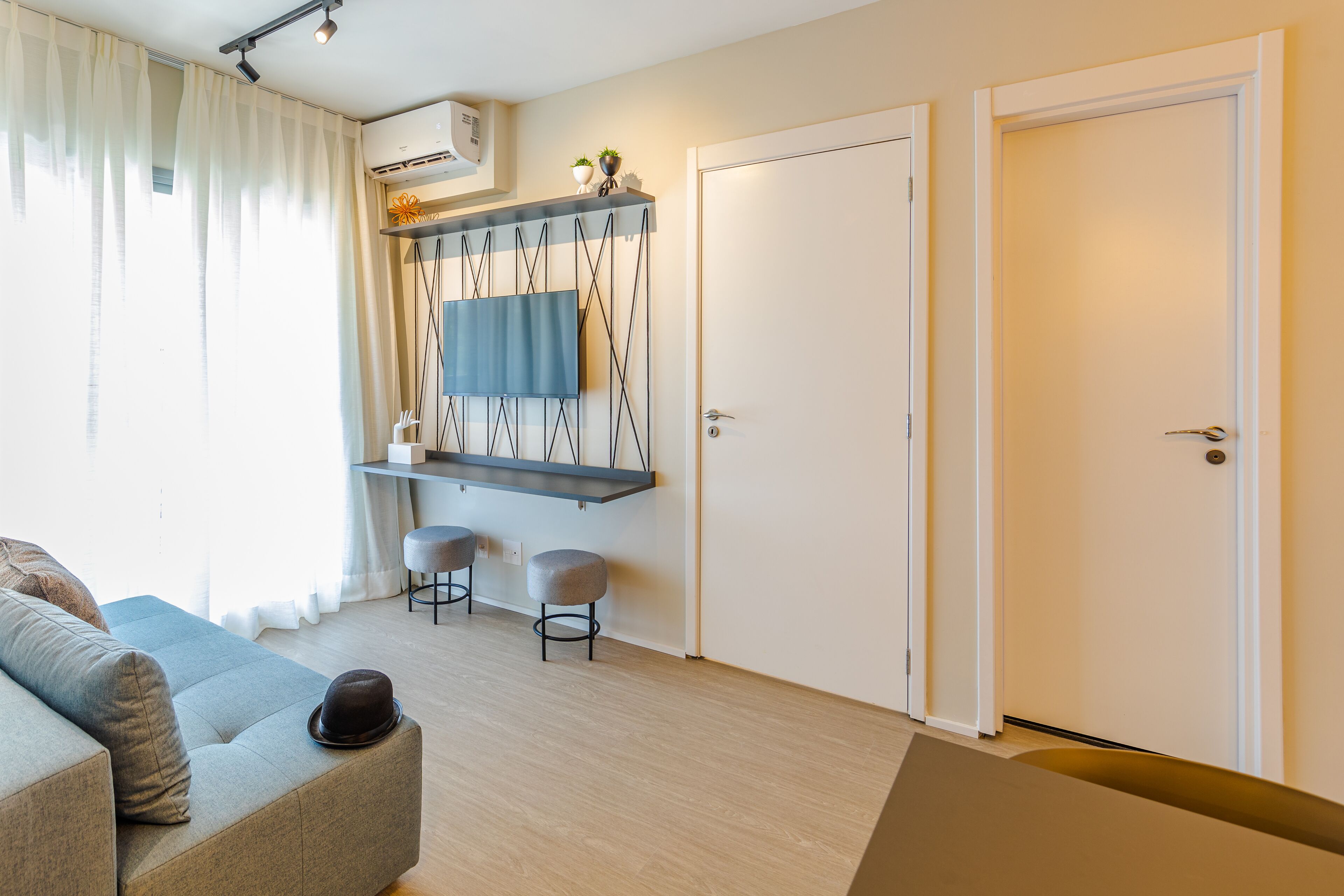 A modern living room with a sofa, a coffee table, and a TV on a wall-mounted shelf. There are two stools and a door in the room.