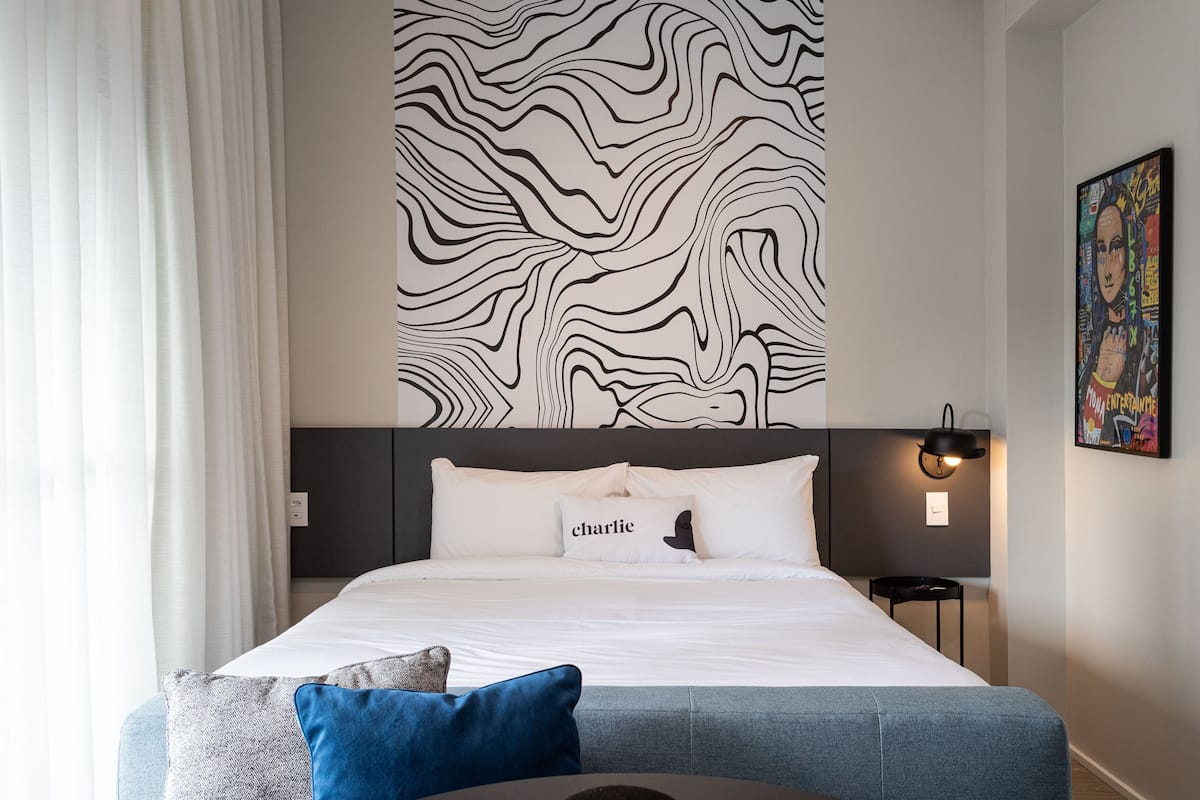Een moderne hotelkamer met een groot bed, een hoofdbord met een golvend patroon, een nachtkastje met een lamp en een kunstwerk aan de muur.