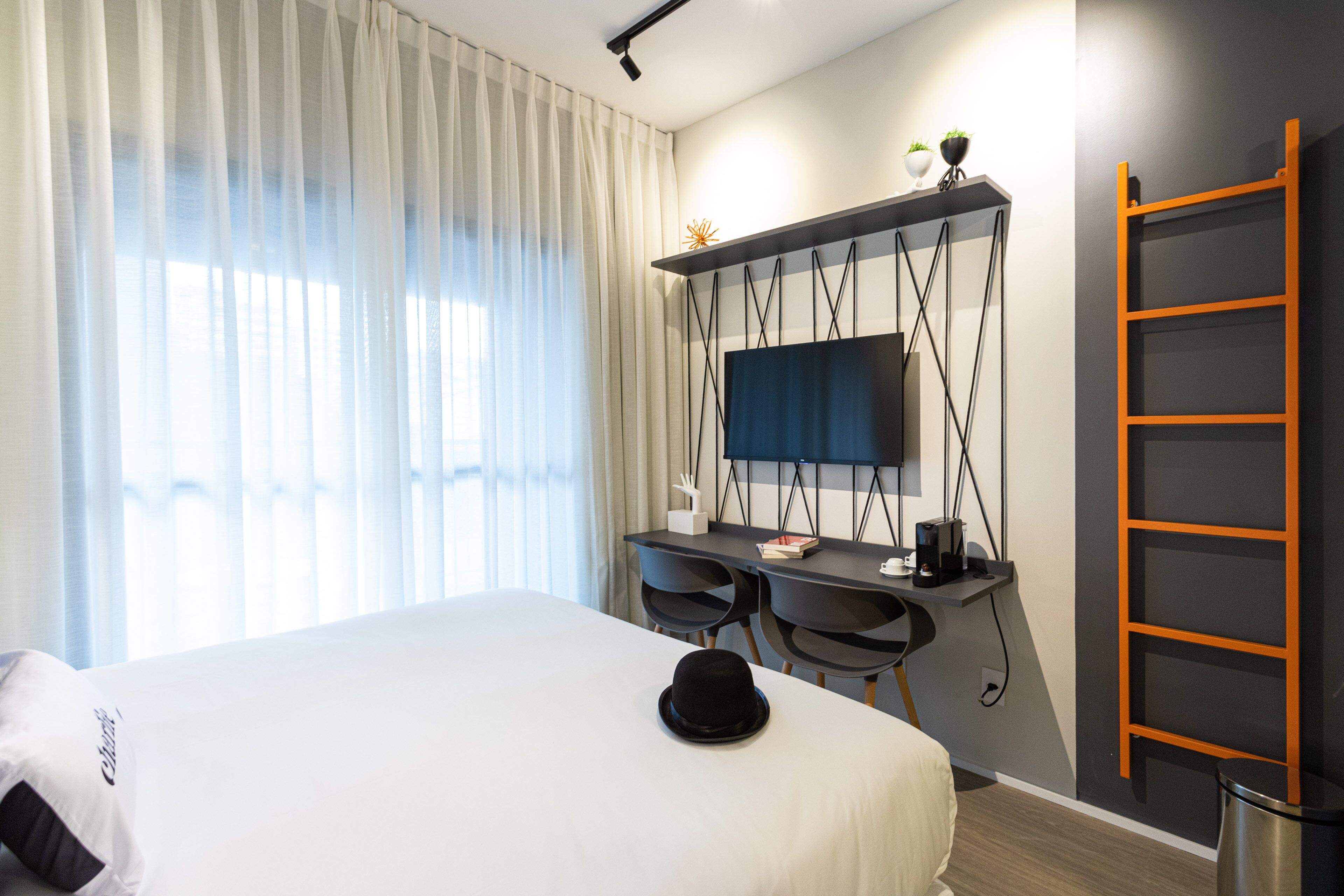 Ein Hotelzimmer mit einem Bett, einem Schreibtisch mit zwei Stühlen, einem Fernseher und einer an der Wand stehenden Leiter.