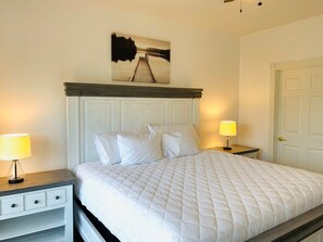 A neatly made bed with white bedding and pillows, a wooden headboard, and a painting above it. There are two bedside tables with lamps on either side of the bed. - Radiant and bright condo in the center of MB (Myrtle Beach)