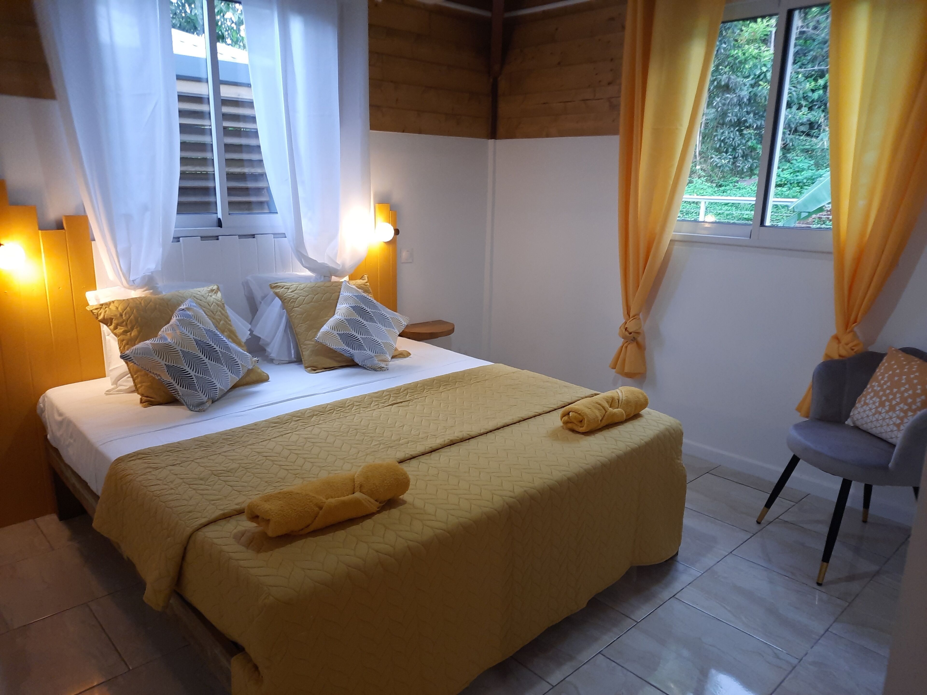 A neatly made bed with a yellow bedspread and pillows, a bedside table with a lamp, and a chair with a cushion.