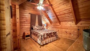 A cozy attic bedroom with wooden walls, a sloped ceiling, a bed with patterned bedding, a nightstand, a lamp, and a ceiling fan. - Labor Day at The Summit - Wifi and Hot Tub! - Callaway Gardens (Warm Springs)