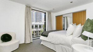 A modern hotel room with a large bed, a small seating area, a nightstand, and a view of the city through a large window.