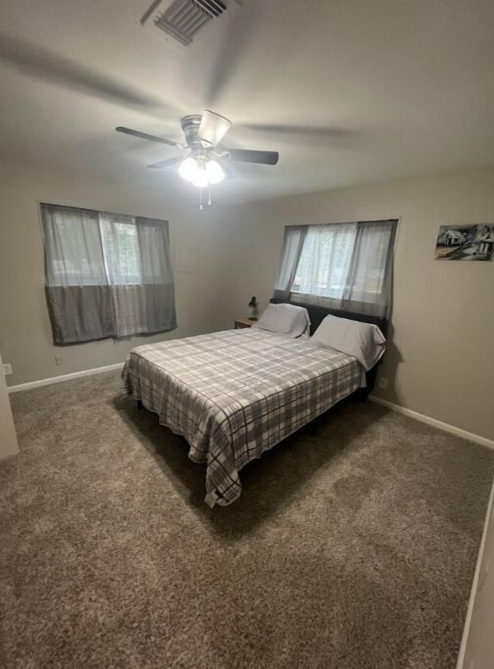 Un dormitorio con una cama, ventilador de techo y dos ventanas con cortinas traslúcidas.