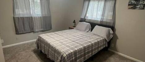 Un dormitorio con una cama, ventilador de techo y dos ventanas con cortinas traslúcidas.