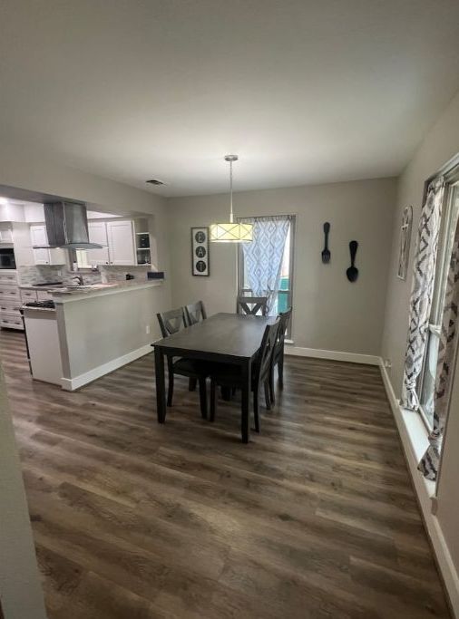 A spacious dining area with a dark wood floor, a central table, and chairs. A kitchen is visible in the background with white cabinets and a hanging light fixture.
