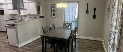 A spacious dining area with a dark wood floor, a central table, and chairs. A kitchen is visible in the background with white cabinets and a hanging light fixture.