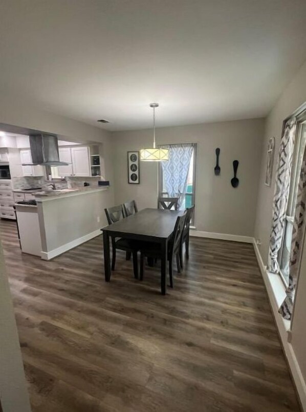 A spacious dining area with a dark wood floor, a central table, and chairs. A kitchen is visible in the background with white cabinets and a hanging light fixture. - Spacious 5BR near Lake Conroe (Montgomery)