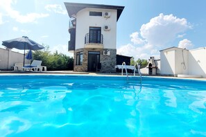 A swimming pool with lounge chairs and an umbrella, surrounded by a modern building with a balcony and stone accents.