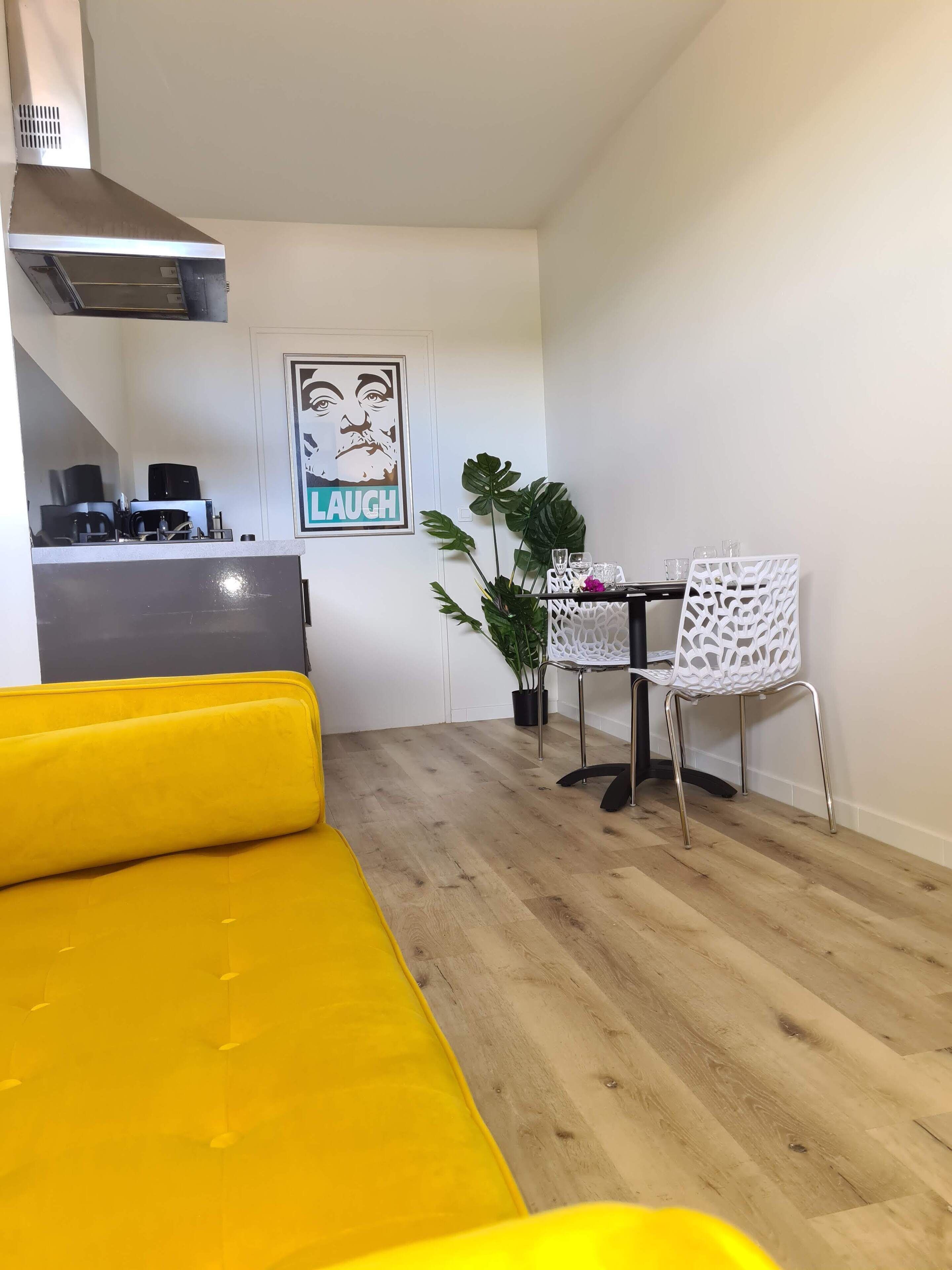 Ein gemütlicher Wohnraum mit einem gelben Sofa, einem Essbereich mit einem weißen Stuhl, einer Küche mit Geräten und einem 'LAUGH'-Poster an der Wand.