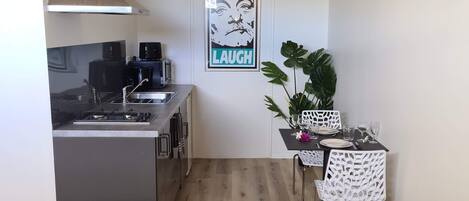 Een compacte keuken met een fornuis, spoelbak en een moderne afzuigkap. Een aangrenzend eetgedeelte met een tafel en stoelen, met een 'LAUGH' poster. Een gele bank is in de voorgrond zichtbaar.