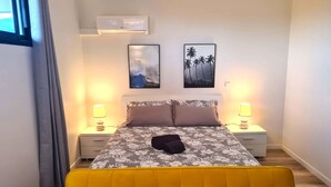 A bedroom with a bed, two bedside tables with lamps, a wall-mounted air conditioner, and two framed pictures. - Studio Ninirei (Papeete)