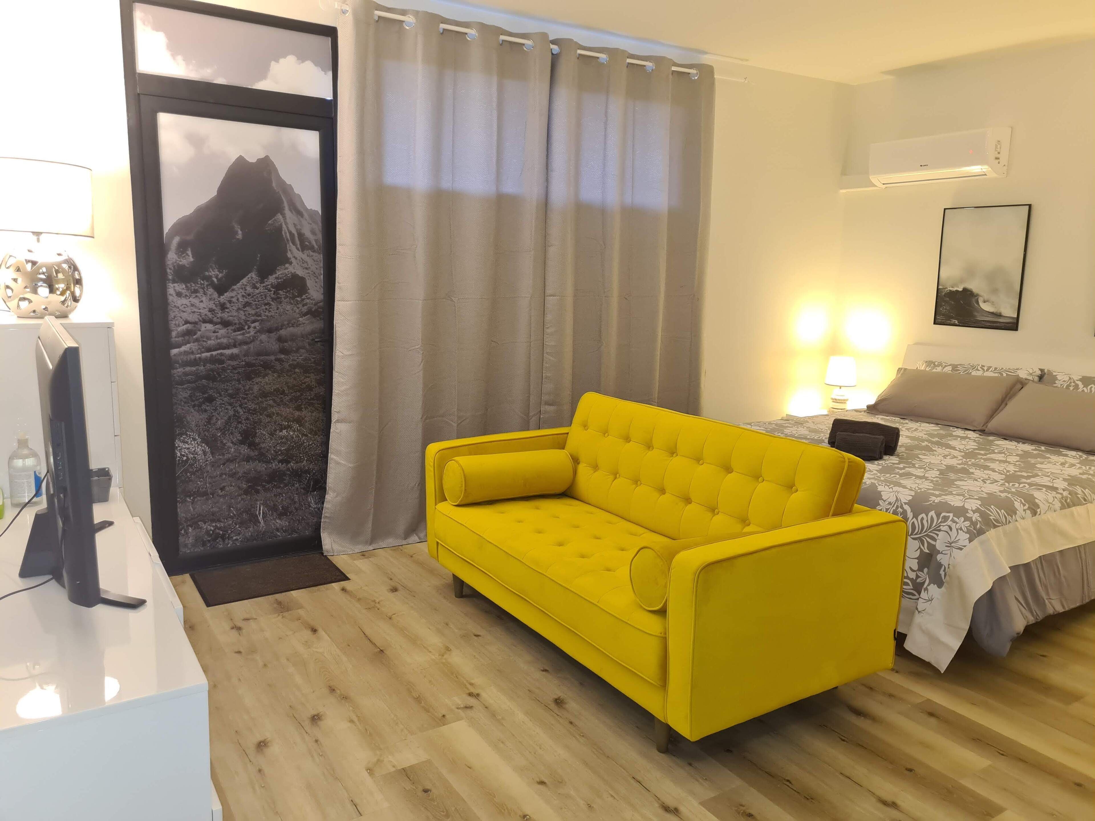 Habitació d'hotel amb un sofà groc, un llit amb roba de llit floral, un aire condicionat penjat i un quadre encimbellat a la paret.