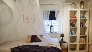 Un dormitorio con cama, mesita de noche, estantería y ventana con vistas a edificios.