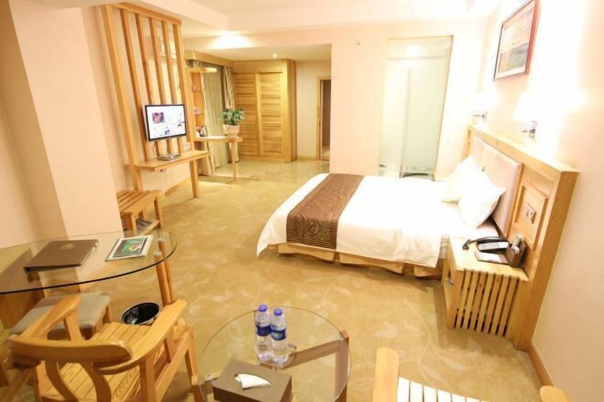 A hotel room with a bed, a desk with a computer, a chair, a small table with a glass top, and a bedside table with a lamp and phone.
