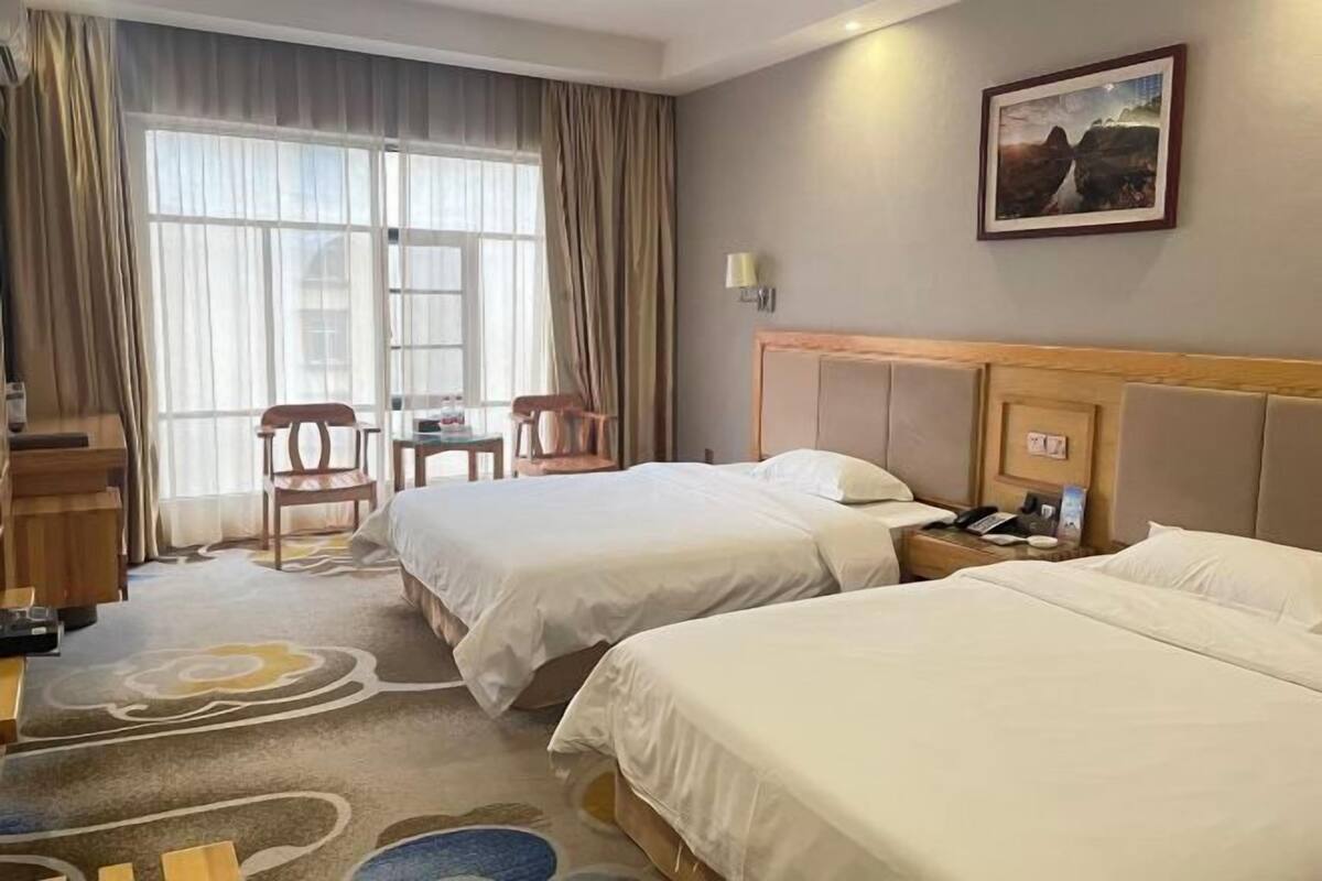 A hotel room with two beds, a desk, chairs, and a large window with curtains.