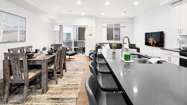 A modern kitchen with a central island, a dining area with wooden chairs, and a living space with a sofa.