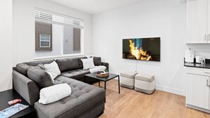 A modern living room with a sectional sofa, a flat-screen TV, a small table, and a kitchen area with cabinets.