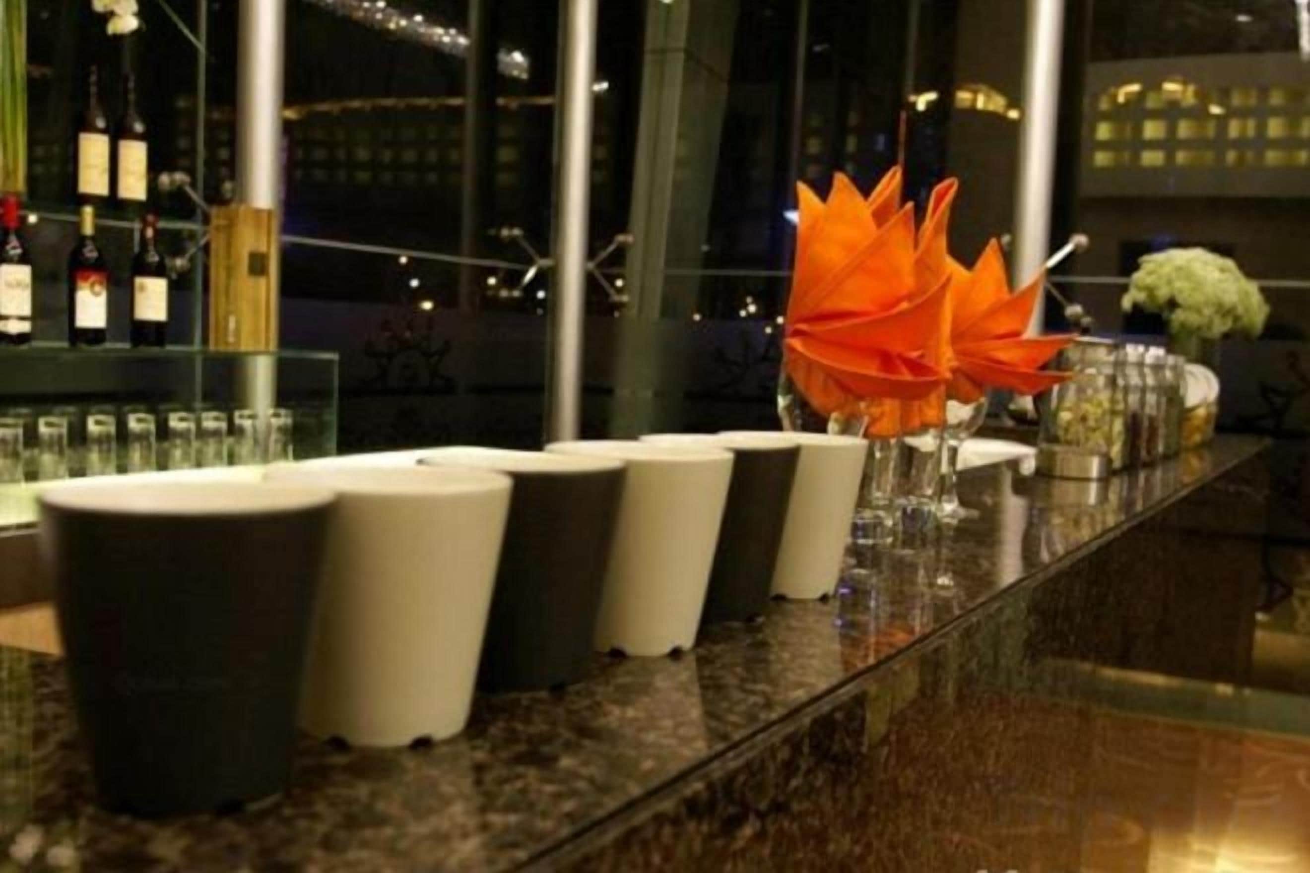 Una zona de bar con una fila de taburetes blanco y negro, una vitrina de vidrio con botellas, y un arreglo floral decorativo de flores naranjas.
