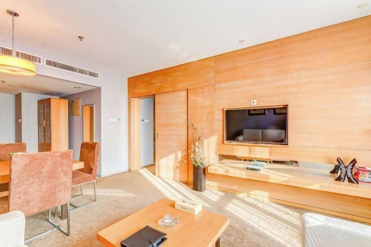A modern hotel room with a wooden panel wall, a flat-screen TV, a sofa, and a dining area with chairs.