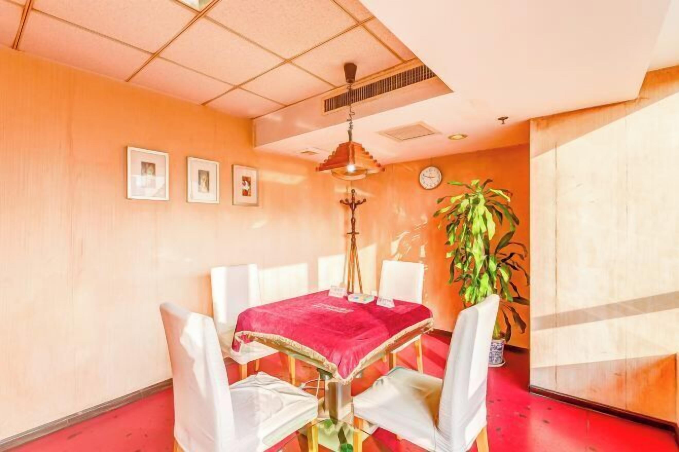 A dining area with a table covered in a red cloth, white chairs, a plant, and framed pictures on the wall.