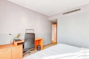 Una habitación de hotel con una cama, un escritorio con una silla, una mesita pequeña, un cubo de basura y una puerta que da a otra habitación.