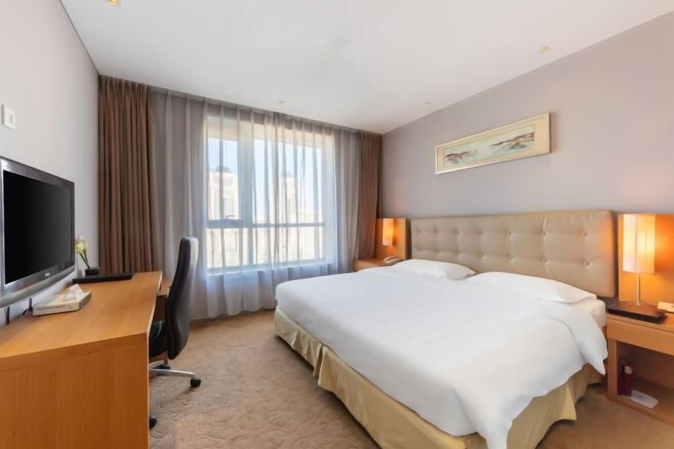 Quarto de hotel com uma cama, uma secretária com televisão, uma cadeira, uma janela com cortinas e um quadro na parede.