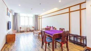 A hotel room with a wooden floor, a dining table with chairs, a sofa, a TV, and a window with curtains.