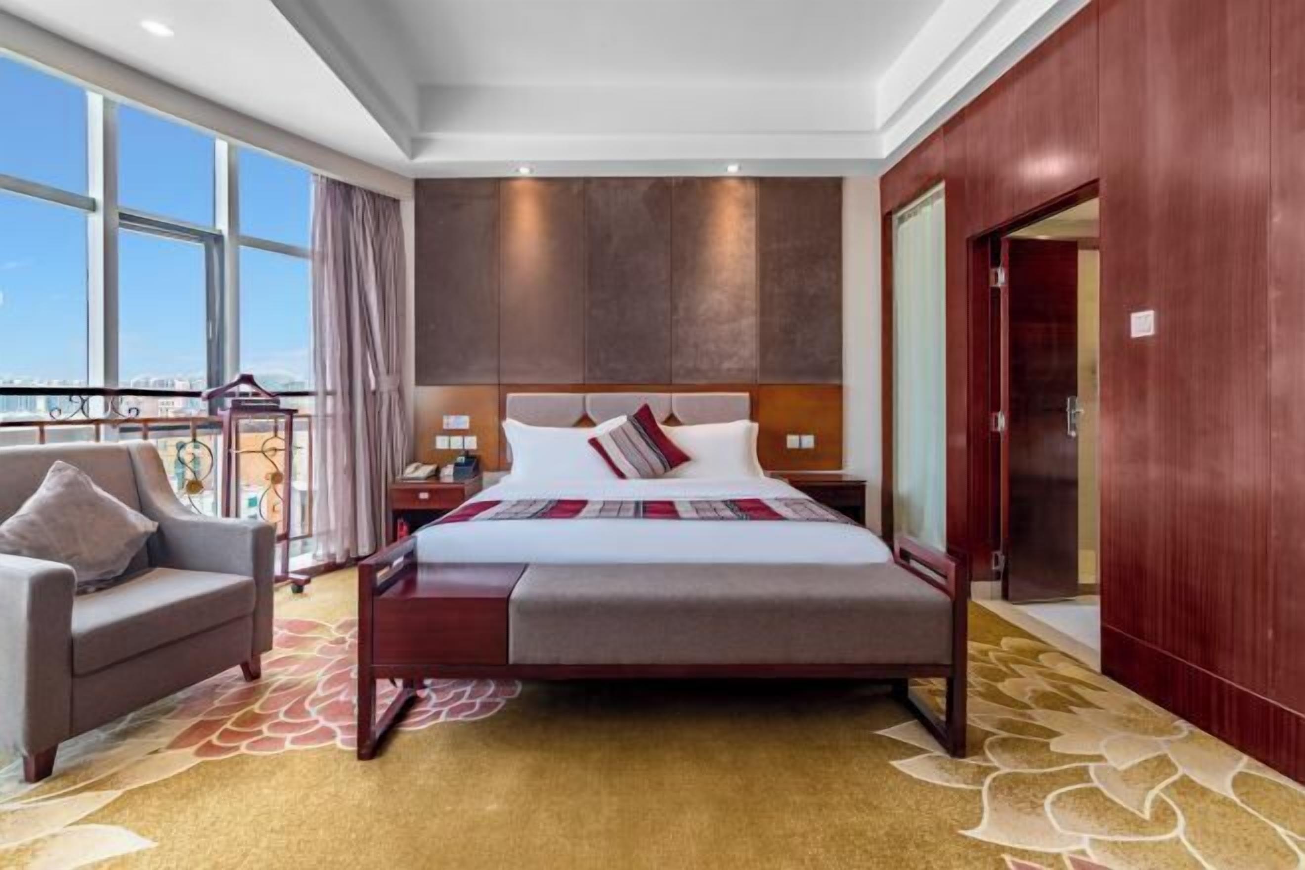 Habitación de hotel con una cama grande, un área de descanso con una silla, una mesita de noche y vistas a la ciudad a través de amplios ventanales.