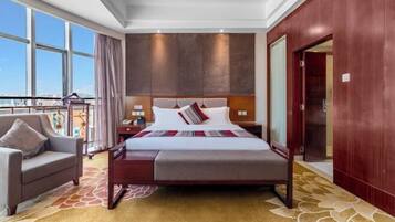 Habitación de hotel con una cama grande, un área de descanso con una silla, una mesita de noche y vistas a la ciudad a través de amplios ventanales.