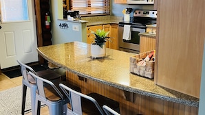 A kitchen with a granite countertop, wooden cabinets, a stainless steel oven, and a microwave. There are three bar stools and a potted plant on the counter. - Cozy Log Cabin on Golf Course, Across from Otsego Lake (Gaylord)