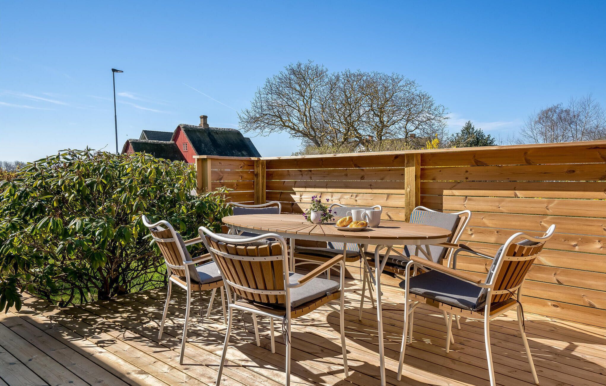 An outdoor wooden deck with a round table and chairs, surrounded by a wooden fence and a hedge.