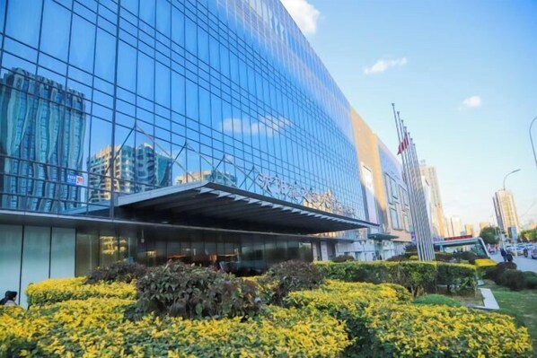 A modern glass building with a reflective facade, surrounded by well-maintained greenery and a clear sky.