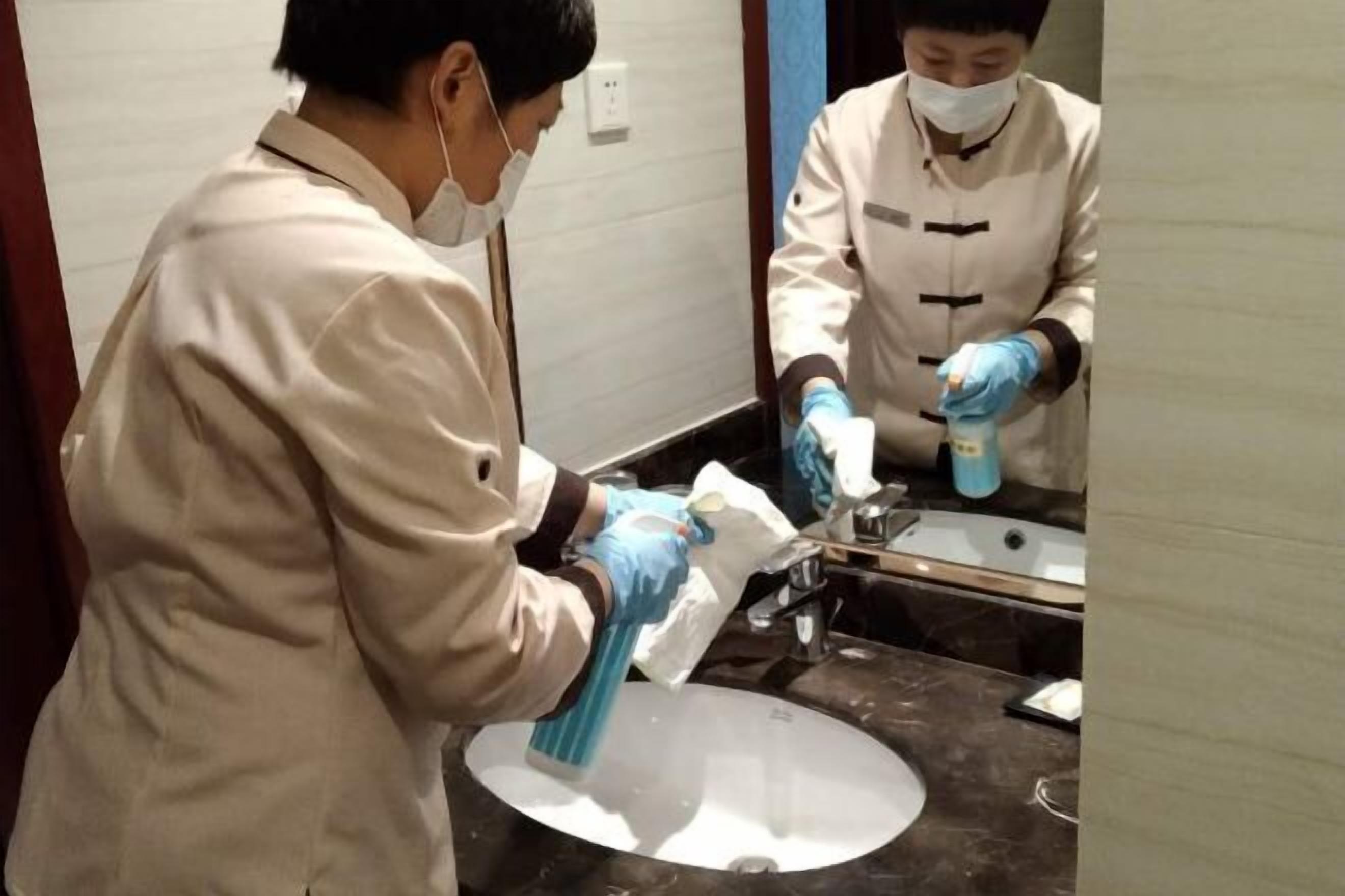 Dos personas limpiando un lavamanos en un baño con un pulverizador y un paño.