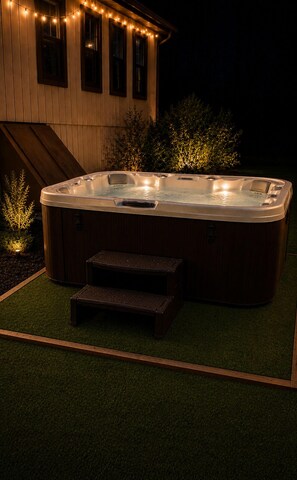 A hot tub with steps, surrounded by artificial grass, against a wooden house with string lights. - Modern Poconos Cabin w/ Hot Tub Near Kalahari & CamelBack Mountain ! (Long Pond)