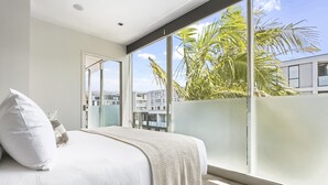 A bedroom with a large window, a bed with white bedding, and a view of buildings and palm trees outside. - Light and convenient city living (Auckland)