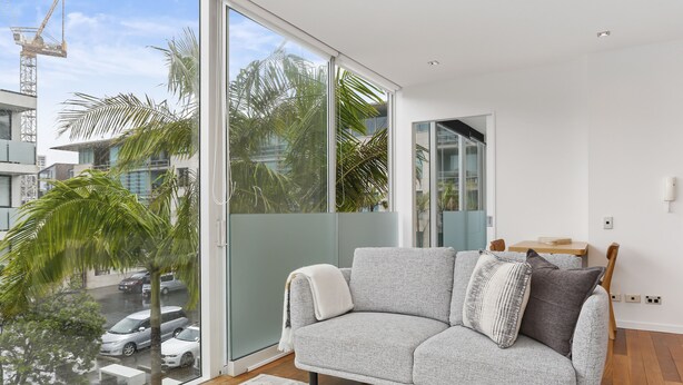 A modern living room with a grey sofa, a round wooden coffee table, and a large sliding glass door leading to a balcony with a view of palm trees and parked cars. - Light and convenient city living (Auckland)