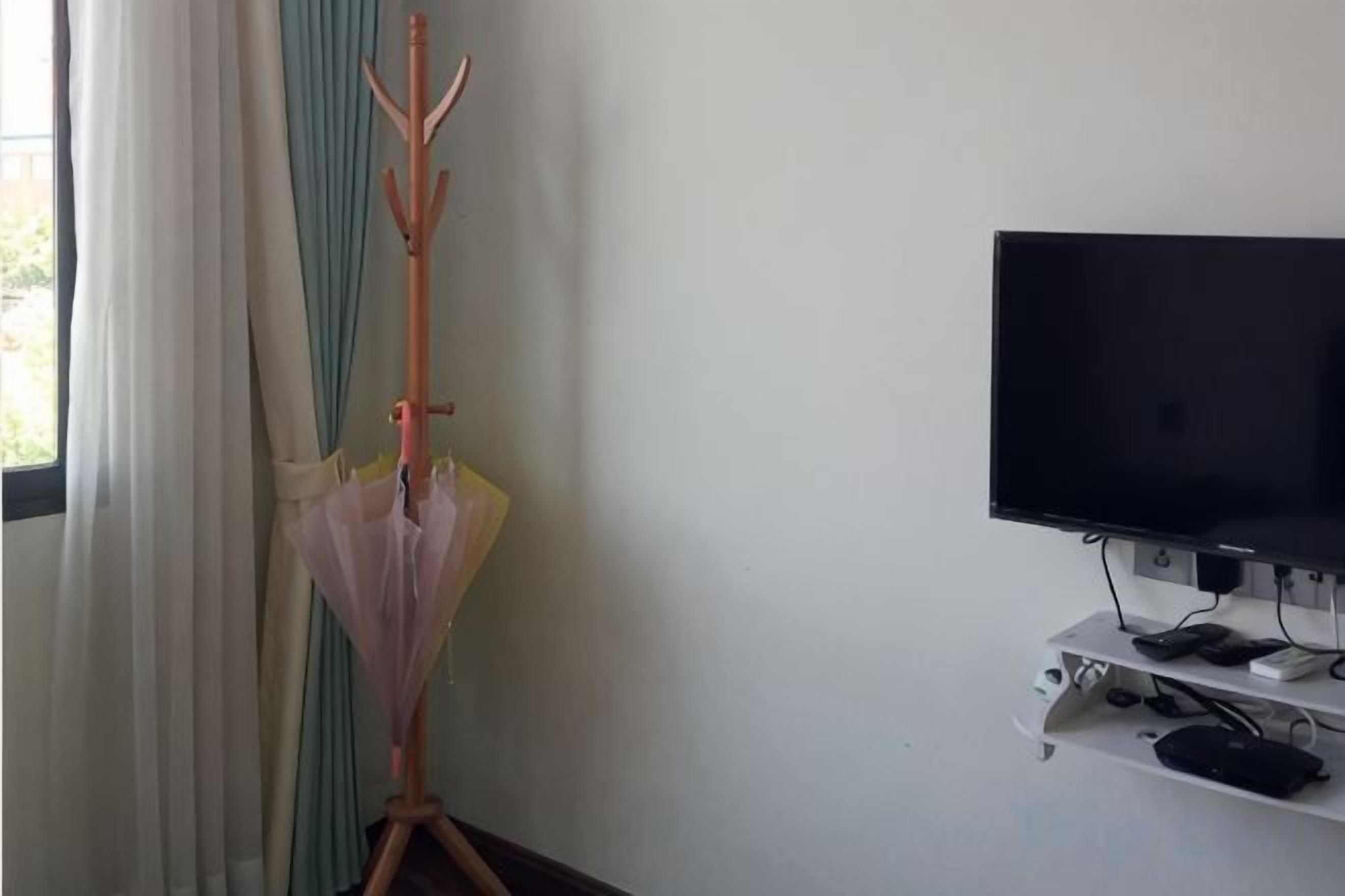 Una habitación con una televisión sobre un mueble bajo, un perchero con un paraguas y una ventana con cortinas.
